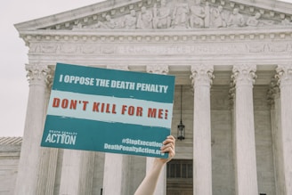 A hand is holding a protest sign in front of a large neoclassical building with columns. The sign reads 'I Oppose the Death Penalty' and 'Don't Kill for Me!', along with hashtags and a website link.