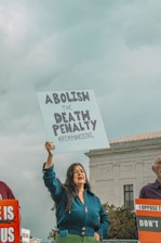 A person holding a sign that reads 'Abolish the Death Penalty' stands outside a government building. The individual appears to be participating in a protest or demonstration. The atmosphere is serious, with overcast skies suggesting a somber mood. Other protesters are partly visible, holding similar signs.