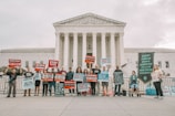 A group of people holding protest signs in front of a large, classical-style government building with columns. The signs carry messages against the death penalty, promoting life and justice. The participants are standing in a single line, displaying various slogans and banners.