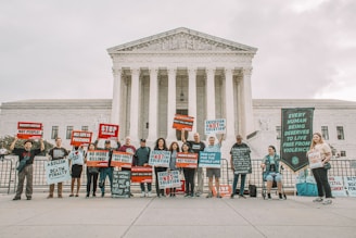 A group of people holding protest signs in front of a large, classical-style government building with columns. The signs carry messages against the death penalty, promoting life and justice. The participants are standing in a single line, displaying various slogans and banners.