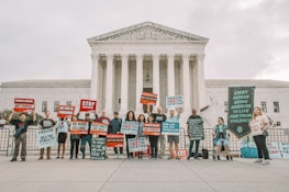 A group of people holding protest signs in front of a large, classical-style government building with columns. The signs carry messages against the death penalty, promoting life and justice. The participants are standing in a single line, displaying various slogans and banners.