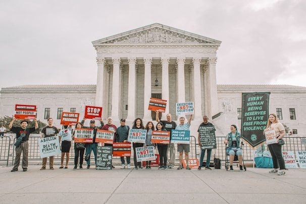 A group of people holding protest signs in front of a large, classical-style government building with columns. The signs carry messages against the death penalty, promoting life and justice. The participants are standing in a single line, displaying various slogans and banners.