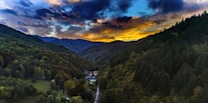 A vibrant sunset illuminates a lush, green valley surrounded by forested mountains. The sky is filled with dramatic clouds painted in hues of orange, yellow, and dark blue. Nestled within the valley are several houses, connected by a narrow road winding through the densely wooded area.