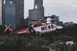 A red and white ambulance helicopter in flight above a cityscape with tall buildings in the background. The helicopter is surrounded by lush green trees at the lower part of the image, adding contrast to the urban setting. The sky is overcast, suggesting a cloudy day.