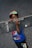 A young child holding a skateboard close to their face, wearing a colorful cap and a blue Spider-Man t-shirt, standing on a textured asphalt surface.