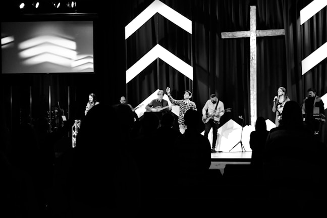 A black and white image of a group of performers on a stage, with musical instruments including guitars and a keyboard. There is a large cross and chevron patterns in the background. Silhouetted figures are visible in the foreground, suggesting an audience.