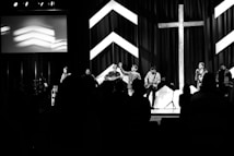 A black and white image of a group of performers on a stage, with musical instruments including guitars and a keyboard. There is a large cross and chevron patterns in the background. Silhouetted figures are visible in the foreground, suggesting an audience.