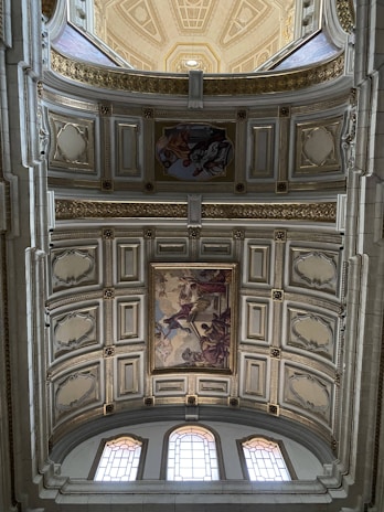 An ornate ceiling with detailed artwork, featuring a series of rectangular panels bordered by golden accents. The central paintings depict classical scenes with human figures. The ceiling architecture includes decorative moldings and arched windows allowing natural light to filter through, highlighting the intricate designs.