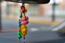 A colorful hanging decoration with various small, cartoon-like animal figures is suspended from a rearview mirror. The background is blurred, indicating an outdoor setting with muted colors.