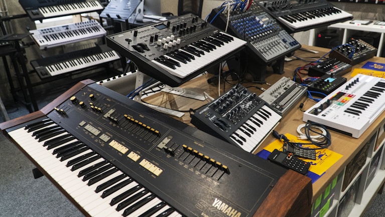 A variety of electronic musical keyboards and synthesizers arranged on a tabletop and stands, including models with an array of dials, switches, and sliders. The setup appears to be in a studio or showroom setting.