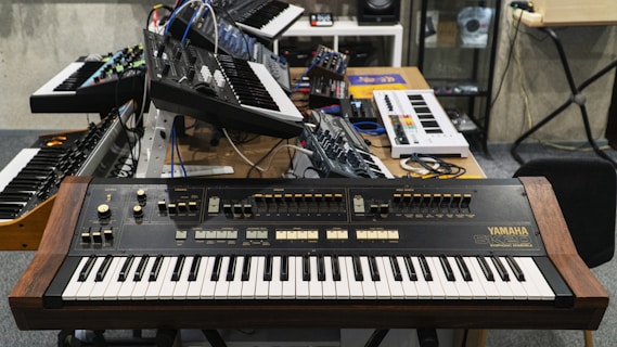 Several synthesizers and keyboards are placed on and around a wooden desk in a studio setting. The main focus is on a Yamaha SY2 synthesizer at the forefront, with various knobs, switches, and keys. Other electronic musical equipment and synthesizers can be seen in the background, with some cables connecting them. The room has a professional, organized appearance suitable for music production.