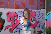 A vibrant graffiti mural on a wall features colorful lettering and a cartoon-style portrait of a woman with long brown hair and green eyes. The background displays a mix of geometric patterns and vibrant colors.