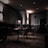 A dimly lit cafe or restaurant interior with a modern design. The room features several tables and wicker chairs, with glass partitions placed between the tables. The walls are decorated with minimalist artwork and warm lighting fixtures. The overall ambiance appears quiet and intimate.