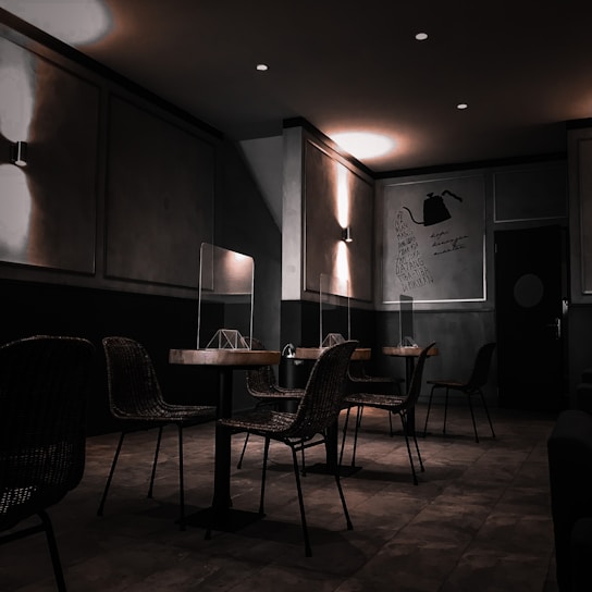 A dimly lit cafe or restaurant interior with a modern design. The room features several tables and wicker chairs, with glass partitions placed between the tables. The walls are decorated with minimalist artwork and warm lighting fixtures. The overall ambiance appears quiet and intimate.