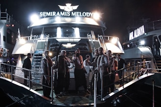 A group of people dressed in matching outfits stands on the deck of a boat named 'Semiramis Melody,' which is illuminated by bright lights. The scene appears to take place at night, with another boat visible in the background with the name 'Ahl Alraya' also lit up.