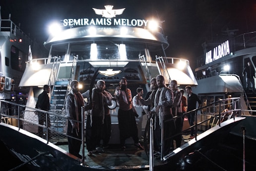 A group of people dressed in matching outfits stands on the deck of a boat named 'Semiramis Melody,' which is illuminated by bright lights. The scene appears to take place at night, with another boat visible in the background with the name 'Ahl Alraya' also lit up.