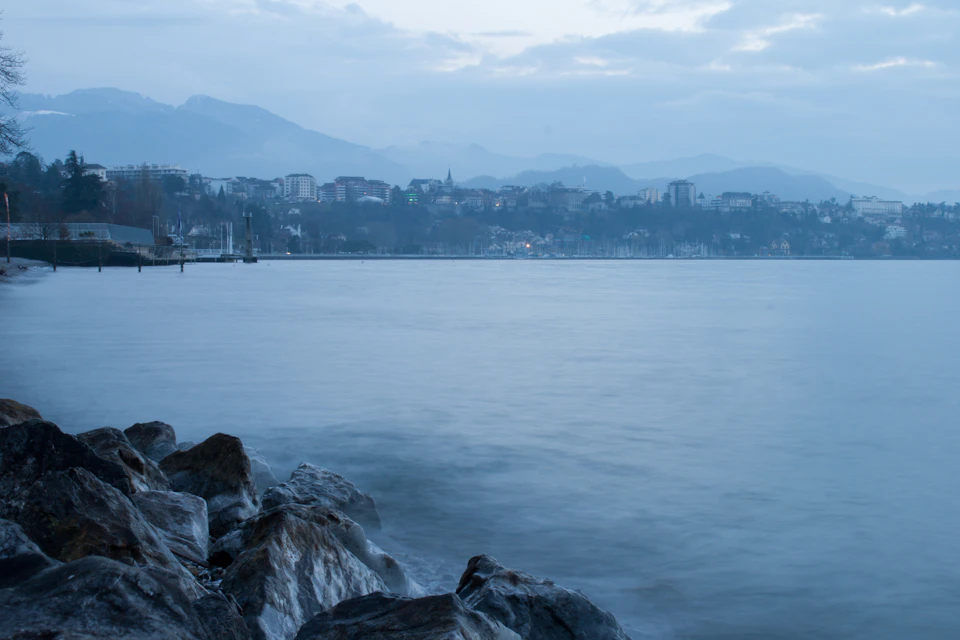 The image depicts a serene lake surrounded by mountains, with a rocky shoreline on one side and a cityscape on the other. The calm water reflects the overcast sky above, creating a tranquil atmosphere. In the distance, several buildings can be seen nestled among the trees, adding depth to the scene. The perspective of the image is from the shore, looking out towards the lake and mountains, providing a comprehensive view of this picturesque landscape.