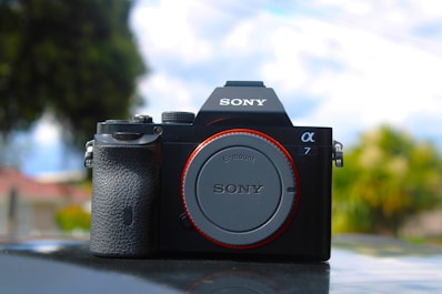 A Sony Alpha 7 camera body, prominently featured against a blurred natural background. The camera is positioned centrally with clear brand and model details visible on its body.