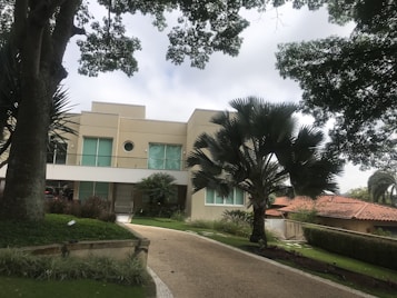 A modern two-story house with a beige facade and large windows is surrounded by lush green trees and a well-maintained garden. A broad driveway leads up to the entrance, flanked by neatly trimmed hedges and decorative plants.
