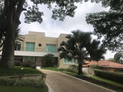 A modern two-story house with a beige facade and large windows is surrounded by lush green trees and a well-maintained garden. A broad driveway leads up to the entrance, flanked by neatly trimmed hedges and decorative plants.