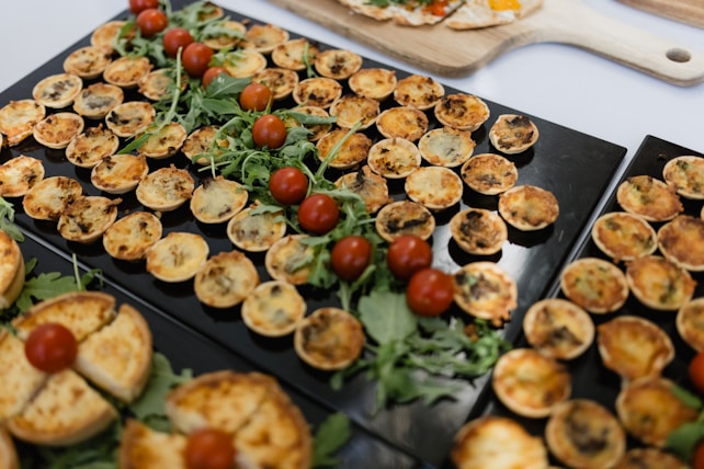A variety of small savory tarts or quiches are arranged on black trays. Each tart appears golden brown with different fillings. There are scattered leafy greens and small cherry tomatoes as garnish.