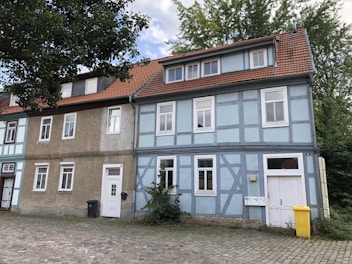 A row of traditional half-timbered houses with light blue and beige facades is seen, featuring red tiled roofs and white-framed windows. The buildings are situated on a cobblestone street, accompanied by a black garbage bin and a yellow recycling bin. Lush green trees are in the background, creating a serene, suburban setting.