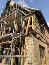 An old building under renovation with wooden beams and scaffolding. The structure has exposed wooden framework and a stone wall section. Scaffolding surrounds the building, indicating ongoing construction work. The sky is visible with a few clouds.