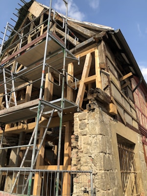 An old building under renovation with wooden beams and scaffolding. The structure has exposed wooden framework and a stone wall section. Scaffolding surrounds the building, indicating ongoing construction work. The sky is visible with a few clouds.