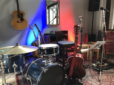 A room contains various musical instruments including a drum set, three acoustic guitars, a microphone, and a music stand with sheet music. The wall behind is lit with blue and red lights. There is a chair next to the drum set, a wooden chair against the wall, and equipment like a speaker and a cable management board on the floor. A reflecting mirror is hung on the wall.