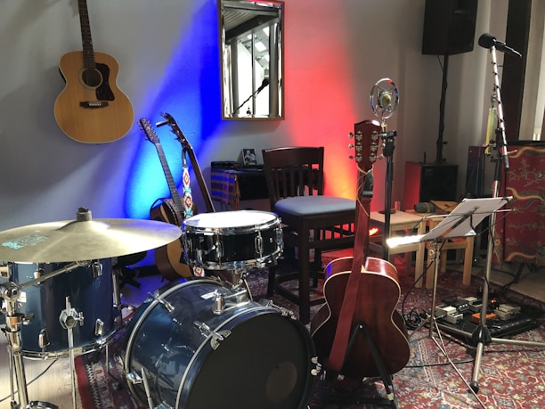 A room contains various musical instruments including a drum set, three acoustic guitars, a microphone, and a music stand with sheet music. The wall behind is lit with blue and red lights. There is a chair next to the drum set, a wooden chair against the wall, and equipment like a speaker and a cable management board on the floor. A reflecting mirror is hung on the wall.