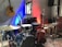 A room contains various musical instruments including a drum set, three acoustic guitars, a microphone, and a music stand with sheet music. The wall behind is lit with blue and red lights. There is a chair next to the drum set, a wooden chair against the wall, and equipment like a speaker and a cable management board on the floor. A reflecting mirror is hung on the wall.