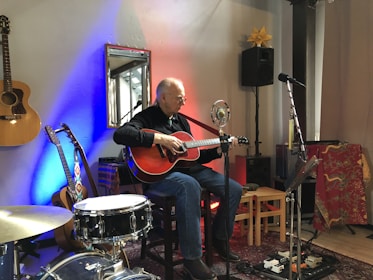 A musician is playing an acoustic guitar on a small stage set up with a microphone and various musical equipment. There are two other guitars visible, one hanging on the wall and another leaning against it. The room has colorful lighting with blue and red hues, and a drum set is partially visible. The setting feels intimate and casual, likely for a small performance or jam session.