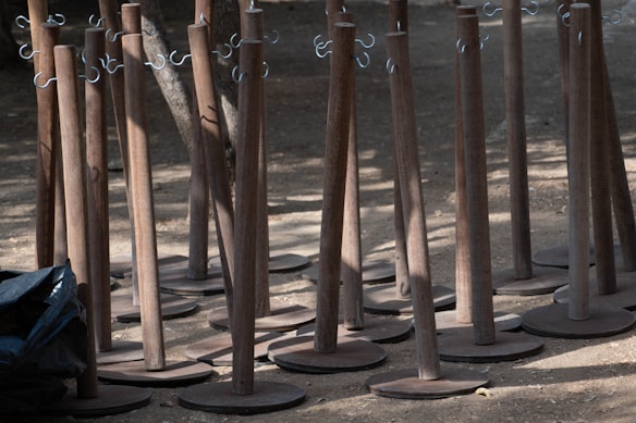 A group of wooden stands with metal hooks are set on a dirt ground. The stands are uniformly arranged, casting shadows in the sunlight. A black plastic bag is partially visible to the side.
