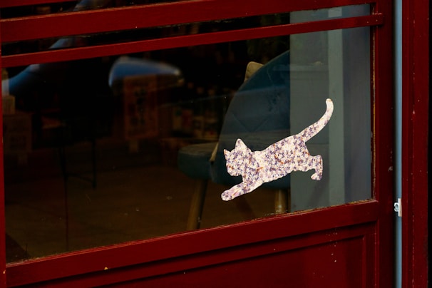 A dark-colored door with a glass pane featuring a decorative sticker of a cat leaping. The interior visible through the glass has elements like chairs and shelves, suggesting a cozy indoor setting.