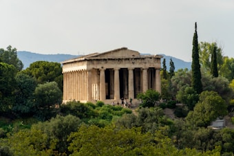 An ancient Greek temple surrounded by lush greenery and trees, set against a backdrop of distant hills and a clear sky. The structure features iconic columns and classical architecture, basking in gentle sunlight with a peaceful ambiance.