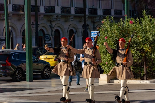 Three soldiers dressed in traditional uniforms with red berets and beige kilts are marching in sync on a city street. They are holding rifles and appear to be part of a ceremonial procession. Behind them, cars are parked, and a building with arched doorways and balconies is visible. Green shrubbery and a few people walking add to the bustling urban scene.
