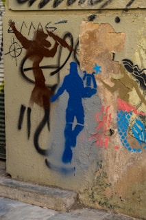 A collection of vibrant graffiti art on a textured wall includes a mix of human silhouettes and abstract patterns. A brown silhouette with outstretched arms stands in front of a nautical wheel, while a blue silhouette holding a hammer or tool is highlighted next to it. There are additional abstract shapes and patterns in red and blue.