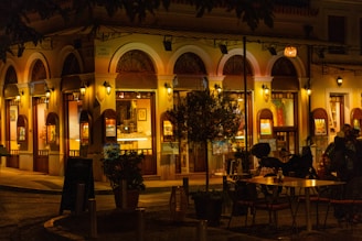 A warmly lit street cafe at night with arched windows and ornate architectural details. Two outdoor tables with chairs are visible, surrounded by potted plants. The interior is illuminated, showing framed artwork on the walls and a cozy ambiance.