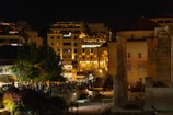 A nighttime cityscape featuring a blend of modern and ancient architecture. There are well-lit buildings and historical ruins, including columns, with a backdrop of residential and commercial structures. The scene is bustling with people, suggesting an active nightlife.