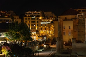 A nighttime cityscape featuring a blend of modern and ancient architecture. There are well-lit buildings and historical ruins, including columns, with a backdrop of residential and commercial structures. The scene is bustling with people, suggesting an active nightlife.