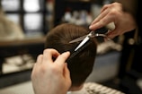 A hairdresser carefully trims a person's short, brown hair using scissors and a comb. The focus is on the precise cutting technique as the hands hold the tools expertly.