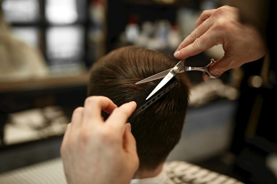 A hairdresser carefully trims a person's short, brown hair using scissors and a comb. The focus is on the precise cutting technique as the hands hold the tools expertly.