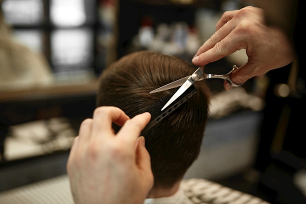 A hairdresser carefully trims a person's short, brown hair using scissors and a comb. The focus is on the precise cutting technique as the hands hold the tools expertly.