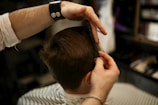 A person with short brown hair is getting a haircut. A barber is holding a comb and using a pair of scissors to trim the hair. The focus is on the barber's hands and the back of the client's head, with a barber shop setting visible in the background.