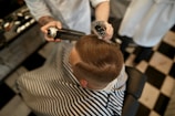 A person is sitting in a barber chair, covered with a striped cape. The barber is styling the person's hair using a round brush and a can of hair spray. The setting appears to be a barbershop with a black and white checkered floor.
