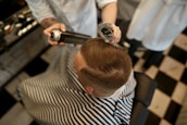 A person is sitting in a barber chair, covered with a striped cape. The barber is styling the person's hair using a round brush and a can of hair spray. The setting appears to be a barbershop with a black and white checkered floor.