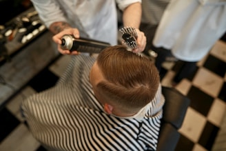 A person is sitting in a barber chair, covered with a striped cape. The barber is styling the person's hair using a round brush and a can of hair spray. The setting appears to be a barbershop with a black and white checkered floor.