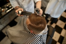 A person is sitting in a barber chair, covered with a striped cape. The barber is styling the person's hair using a round brush and a can of hair spray. The setting appears to be a barbershop with a black and white checkered floor.
