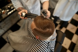 A person is sitting in a barber chair, covered with a striped cape. The barber is styling the person's hair using a round brush and a can of hair spray. The setting appears to be a barbershop with a black and white checkered floor.