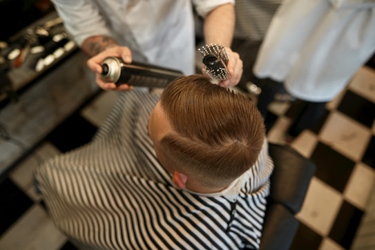 A person is sitting in a barber chair, covered with a striped cape. The barber is styling the person's hair using a round brush and a can of hair spray. The setting appears to be a barbershop with a black and white checkered floor.